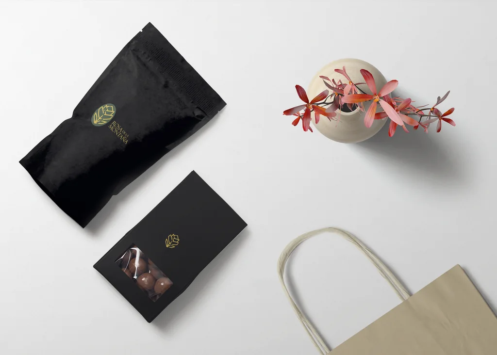 Empaques negros de café y chocolate, con bolsa de papel y flores sobre un fondo claro.